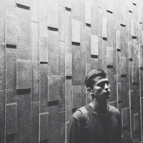 A monochrome image of a man dressed in black, standing confidently against a metallic textured wall.