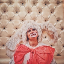 A person is dressed in a costume resembling a rabbit or a fluffy creature, complete with a headpiece featuring long, drooping ears and face paint including whiskers and a red nose. The costume includes a large, bright red bow. The background is a padded, tufted surface with buttons in various colors.