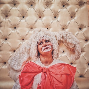 A person is dressed in a costume resembling a rabbit or a fluffy creature, complete with a headpiece featuring long, drooping ears and face paint including whiskers and a red nose. The costume includes a large, bright red bow. The background is a padded, tufted surface with buttons in various colors.