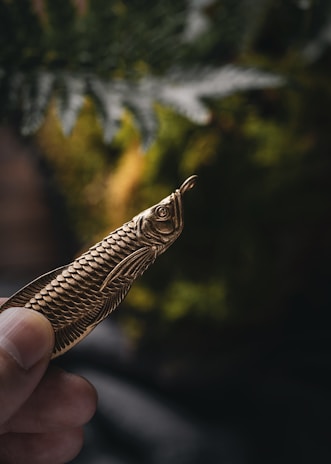 A hand is shown holding a brass object shaped like a fish with detailed scale patterns. The background is blurred with hints of green foliage.