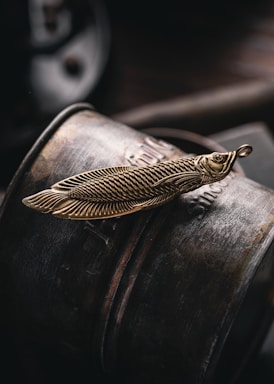 A detailed view of a brass object shaped like a feather with intricate patterns, resting on a rounded, metallic, textured surface. The lighting is soft, creating shadows and highlights that emphasize the object's design.