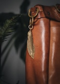 A close-up view of a brown leather bag featuring a decorative metal fish-shaped charm hanging from a brass ring. The background includes green plant leaves, adding a natural element to the scene.