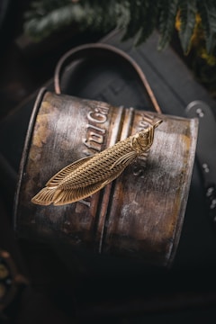 A metal object with intricate engravings resembling a fish is placed on a curved, rustic, cylindrical surface, possibly a barrel or canister. The object appears gold in color with detailed scales and a textured finish. The background hints at possible greenery and a dark, rich ambiance.