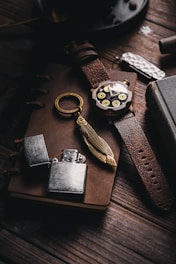 A rustic arrangement featuring a leather-bound notebook, a vintage wristwatch with brass details, an ornate keychain with a feather charm, a metal lighter, and a textured pocket knife. The items rest on a wooden surface, creating an antique and nostalgic ambiance.