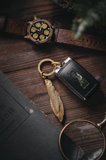 A close-up view features a textured wooden surface on which several items are arranged. A leather-strapped watch with a unique design resembling bullet casings is placed near a key fob adorned with a fish-shaped metallic keychain. A portion of a vintage-looking book and a pair of glasses lie nearby, adding to the rustic and sophisticated atmosphere.