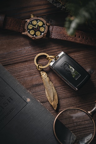 A close-up view features a textured wooden surface on which several items are arranged. A leather-strapped watch with a unique design resembling bullet casings is placed near a key fob adorned with a fish-shaped metallic keychain. A portion of a vintage-looking book and a pair of glasses lie nearby, adding to the rustic and sophisticated atmosphere.