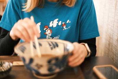 A person is seated at a table holding chopsticks above a large ceramic bowl. The individual is wearing a blue shirt featuring a design of two figures skiing on a snowy landscape. The atmosphere appears casual and relaxed, suggesting a meal setting, possibly in a restaurant.