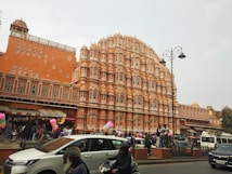 A large, intricately designed pink sandstone building with numerous small windows and a detailed facade. The architecture features archways and small pavilions, typical of traditional Indian design. In front of the building, there is a bustling scene with people walking and gathering, as well as several parked and moving vehicles. Some street vendors are visible along the side of the street, with colorful balloons and other merchandise displayed.