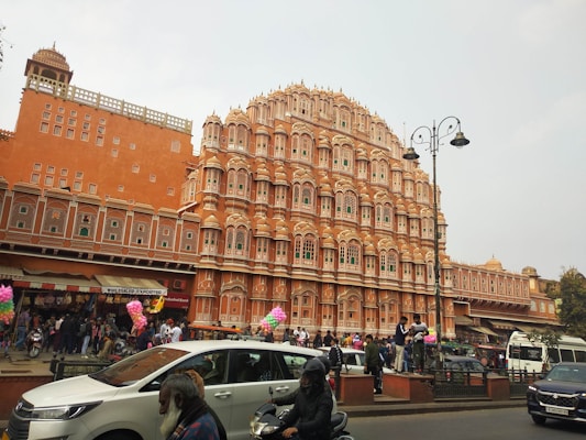A large, intricately designed pink sandstone building with numerous small windows and a detailed facade. The architecture features archways and small pavilions, typical of traditional Indian design. In front of the building, there is a bustling scene with people walking and gathering, as well as several parked and moving vehicles. Some street vendors are visible along the side of the street, with colorful balloons and other merchandise displayed.