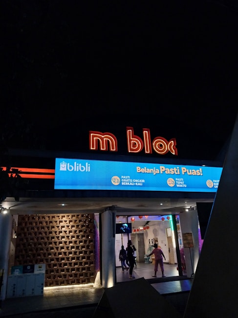 An illuminated sign of 'm bloc' is displayed prominently with a blue storefront banner below advertising 'Belanja Pasti Puas!' from a company called Blibli. The entrance shows a modern design with geometric patterns on the wall and a few people walking inside under a ceiling with hanging decorations.