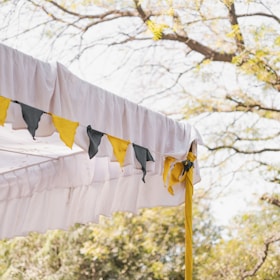 A white fabric tent with decorative bunting in yellow and green hanging from the edge. The tent is set outdoors, visible through tree branches with fresh green leaves under a bright sky.