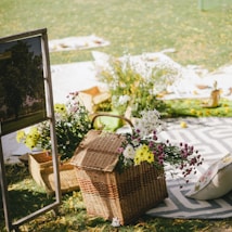 A picturesque outdoor picnic arrangement featuring a wicker basket filled with colorful flowers, including white, yellow, and purple blooms. The setup includes patterned blankets, cushions, and a sign with a tree image. The setting is bright and green, with sunlight casting gentle shadows.