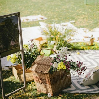 A picturesque outdoor picnic arrangement featuring a wicker basket filled with colorful flowers, including white, yellow, and purple blooms. The setup includes patterned blankets, cushions, and a sign with a tree image. The setting is bright and green, with sunlight casting gentle shadows.
