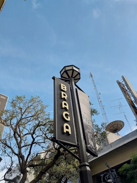 A street sign displaying the word 'Braga' is prominently featured, with a clear blue sky in the background. There are trees partially visible around the sign, and antennas are seen on top of nearby buildings.
