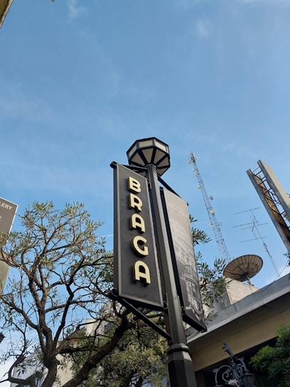A street sign displaying the word 'Braga' is prominently featured, with a clear blue sky in the background. There are trees partially visible around the sign, and antennas are seen on top of nearby buildings.