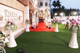A beautifully decorated outdoor banquet hall with a red carpet leading to an ornate entryway. The area is adorned with floral arrangements set atop tall, white vases. There are tables and people in the background under soft lighting, and the setting suggests a celebration or event.