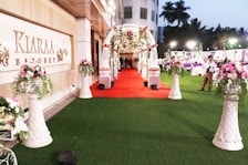 A beautifully decorated outdoor banquet hall with a red carpet leading to an ornate entryway. The area is adorned with floral arrangements set atop tall, white vases. There are tables and people in the background under soft lighting, and the setting suggests a celebration or event.