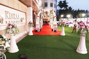 A beautifully decorated outdoor banquet hall with a red carpet leading to an ornate entryway. The area is adorned with floral arrangements set atop tall, white vases. There are tables and people in the background under soft lighting, and the setting suggests a celebration or event.