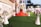 A beautifully decorated outdoor banquet hall with a red carpet leading to an ornate entryway. The area is adorned with floral arrangements set atop tall, white vases. There are tables and people in the background under soft lighting, and the setting suggests a celebration or event.
