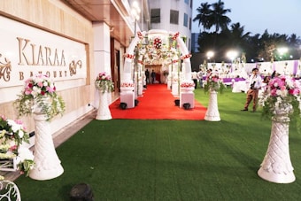 A beautifully decorated outdoor banquet hall with a red carpet leading to an ornate entryway. The area is adorned with floral arrangements set atop tall, white vases. There are tables and people in the background under soft lighting, and the setting suggests a celebration or event.