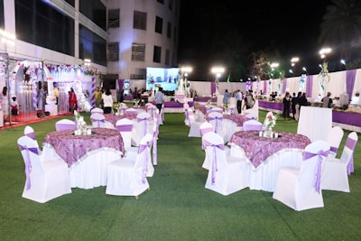 A well-decorated outdoor event space is set up with round tables covered in purple and white tablecloths and chairs wrapped with white covers and purple bows. Floral centerpieces are placed on the tables. The venue is adorned with white and purple drapes, and there are decorative lights illuminating the area. In the background, people are mingling near a buffet section. The scene is lively and festive.
