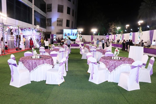 A well-decorated outdoor event space is set up with round tables covered in purple and white tablecloths and chairs wrapped with white covers and purple bows. Floral centerpieces are placed on the tables. The venue is adorned with white and purple drapes, and there are decorative lights illuminating the area. In the background, people are mingling near a buffet section. The scene is lively and festive.