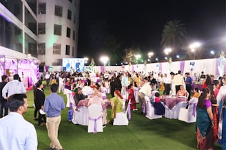 A large outdoor gathering with people dressed in formal and traditional attire, seated at tables on a grass-covered area. The setting includes colorful decorations, lighting, and a large screen displaying an image in the background.