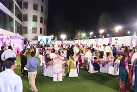 A large outdoor gathering with people dressed in formal and traditional attire, seated at tables on a grass-covered area. The setting includes colorful decorations, lighting, and a large screen displaying an image in the background.