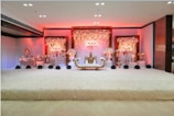 An elegant and luxurious stage setup featuring a lavishly decorated sofa in the center, surrounded by ornate floral arrangements. The backdrop is adorned with cascading flowers and soft, ambient lighting that emits a warm glow. The overall decor is rich with intricate details, creating an opulent and festive atmosphere.