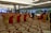 A large, elegantly decorated banquet hall with rows of red and gold chairs arranged for an event. People are mingling near tables set for dining. Chandeliers and lighting add a luxurious ambiance, and the floor features a patterned carpet.