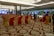 A large, elegantly decorated banquet hall with rows of red and gold chairs arranged for an event. People are mingling near tables set for dining. Chandeliers and lighting add a luxurious ambiance, and the floor features a patterned carpet.