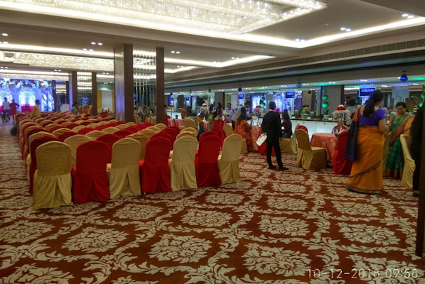 A large, elegantly decorated banquet hall with rows of red and gold chairs arranged for an event. People are mingling near tables set for dining. Chandeliers and lighting add a luxurious ambiance, and the floor features a patterned carpet.