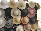A collection of wide-brimmed hats displayed on a wall. The hats vary in color, including shades of cream, black, and pink. Some are adorned with decorative elements like ribbons and beads.