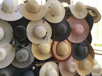 A collection of wide-brimmed hats displayed on a wall. The hats vary in color, including shades of cream, black, and pink. Some are adorned with decorative elements like ribbons and beads.