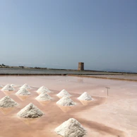Several neatly arranged piles of white salt are drying on a pinkish salt flat. In the background, there is a large water body and an old stone tower. The sky is clear with a hint of haze, suggesting a sunny day.