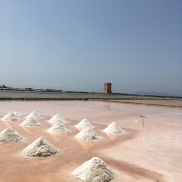 Several neatly arranged piles of white salt are drying on a pinkish salt flat. In the background, there is a large water body and an old stone tower. The sky is clear with a hint of haze, suggesting a sunny day.