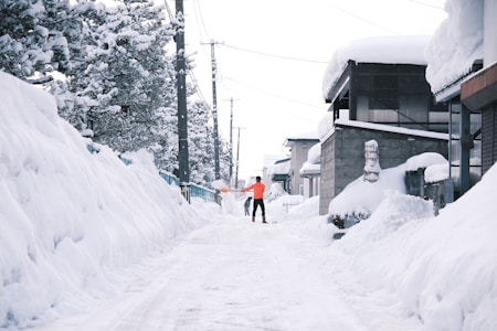 A snowy neighborhood scene with tall snowbanks lining both sides of a narrow street. A person wearing a bright orange jacket is shoveling snow, surrounded by houses covered with thick layers of snow. Snow-laden trees and utility poles are visible, adding to the wintry atmosphere.