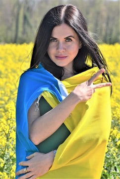 A woman stands in a field of yellow flowers, wrapped in a blue and yellow flag. She is making a peace sign with her fingers and gazes confidently at the camera. Her black hair cascades down her shoulders as she stands against a blurred background of wildflowers and trees.
