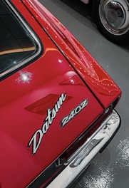 Close-up view of a red car featuring the Datsun 240Z badge. The car's sleek lines and shiny surface indicate its well-maintained condition. Reflections and ambient lighting add to its aesthetic appeal.