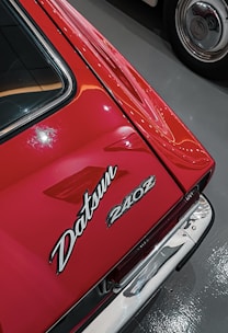 Close-up view of a red car featuring the Datsun 240Z badge. The car's sleek lines and shiny surface indicate its well-maintained condition. Reflections and ambient lighting add to its aesthetic appeal.