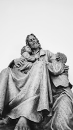 A statue depicting a seated figure with two children resting against him. The central figure appears to be an adult with long hair and a beard, exuding a sense of calm and compassion. One child is nestled on his lap, while the other leans affectionately against his side. The sculpture is monochromatic, highlighting intricate details in the clothing and expressions.