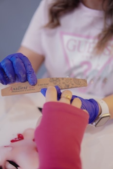 A person wearing blue gloves is holding a nail file labeled 'Analiese Linx Professional Cosmetics.' Another person with a pink sleeve and visible wristwatch is reaching out, suggesting a manicure or nail care session. The background displays a partially visible graphic or logo on a t-shirt.