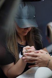 A person wearing a black cap and black shirt is focused on a detailed task, carefully painting or decorating nails. The setting suggests a nail salon environment with tools visible.