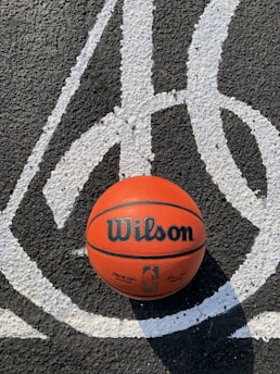 A basketball sits on an asphalt surface with white painted lines. The ball has 'Wilson' and an NBA logo printed on it. Shadows are visible, indicating a sunny day.