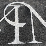 A section of textured black asphalt with large, white-painted letters. The letters have a serif style and a slightly worn-out appearance.