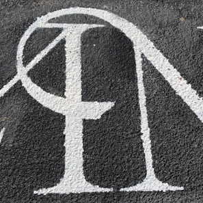 A section of textured black asphalt with large, white-painted letters. The letters have a serif style and a slightly worn-out appearance.