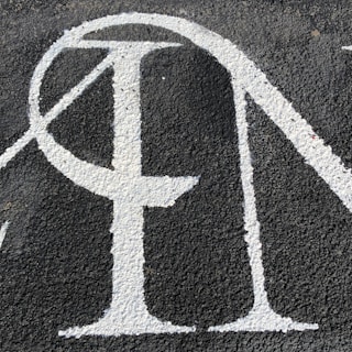 A section of textured black asphalt with large, white-painted letters. The letters have a serif style and a slightly worn-out appearance.