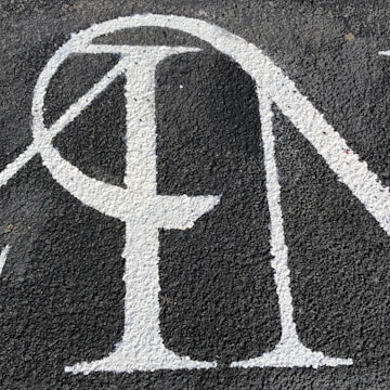 A section of textured black asphalt with large, white-painted letters. The letters have a serif style and a slightly worn-out appearance.