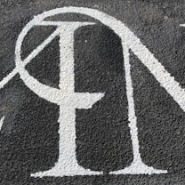 A section of textured black asphalt with large, white-painted letters. The letters have a serif style and a slightly worn-out appearance.