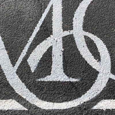 A detailed close-up of a stylized white letter 'A' painted on a rough, dark asphalt surface. The paint appears slightly chipped and worn, with noticeable texture due to the underlying surface.
